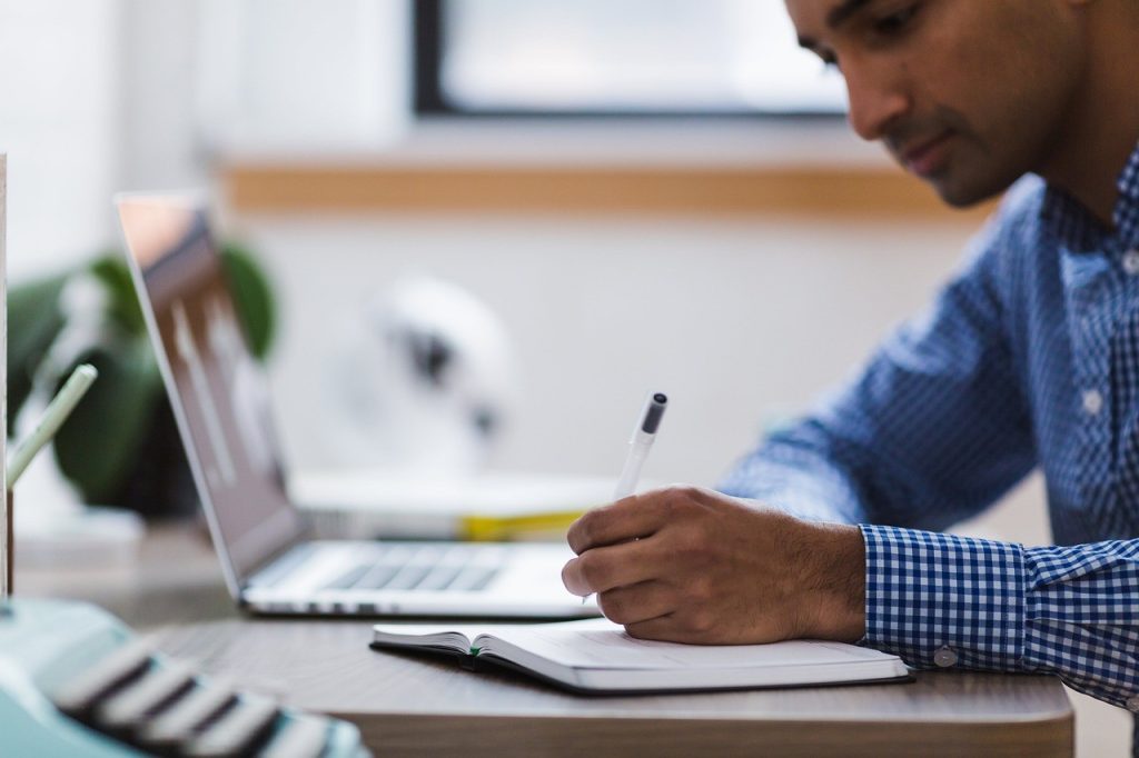A man takes handwritten notes beside a laptop in a bright, plant-filled office